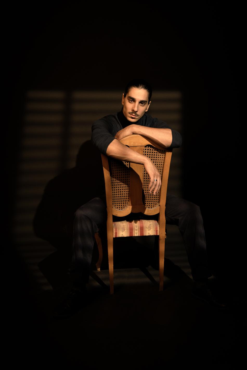 Actor seated backwards on a cane chair, arms folded on the rail