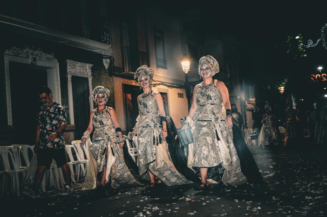 Three women in silver costume walking down a lamplit street