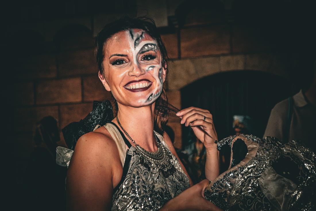 Woman smiling widely in ornate face paint against a brick wall
